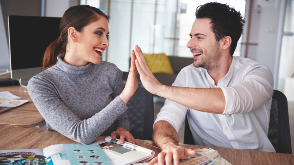 A man and a woman are sitting at a desk, smiling and giving each other a high five. The woman is wearing a grey sweater and the man is wearing a white shirt. They are both looking at each other and appear to be happy and excited. There are papers and magazines on the desk in front of them. This image represents a moment of celebration or success. The high five symbolizes teamwork, collaboration, and shared accomplishment. The smiles on their faces suggest that they are enjoying their work and their relationship. The image captures a positive and optimistic atmosphere.