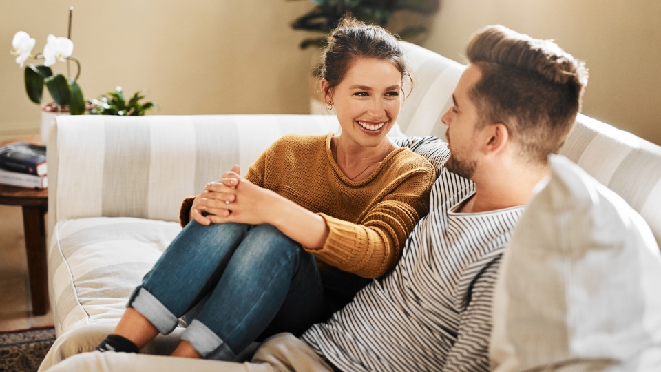 A couple is sitting on a white couch. The woman is wearing a mustard yellow sweater and blue jeans. She is looking at the man with a smile on her face. The man is wearing a striped shirt and has a beard. He is looking at the woman with a smile on his face. The couple is sitting close together, and it appears that they are in love. The couch is in a well-lit room with plants in the background.